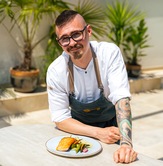 Un chef portant un tablier est appuyé contre une table, près d’une assiette de saumon et de légumes 