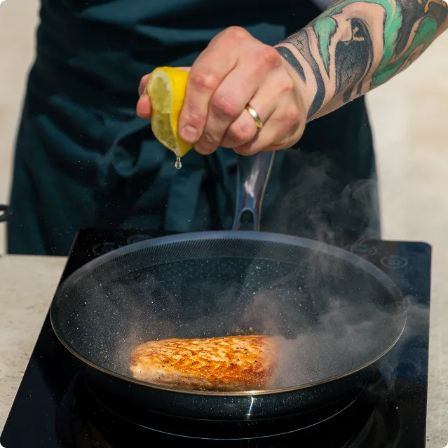Détail d’une main pressant un citron dans une poêle avec du saumon en préparation